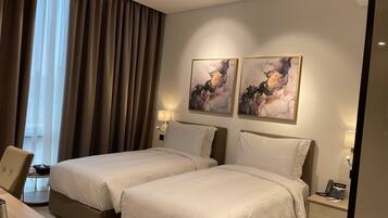 A hotel room with two beds, a desk with a chair, a bedside table with a lamp, and artwork on the wall.