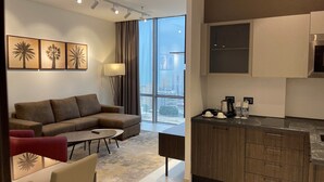 A modern hotel room with a sofa, a coffee table, a dining table, and a kitchen area with cabinets and a coffee machine.