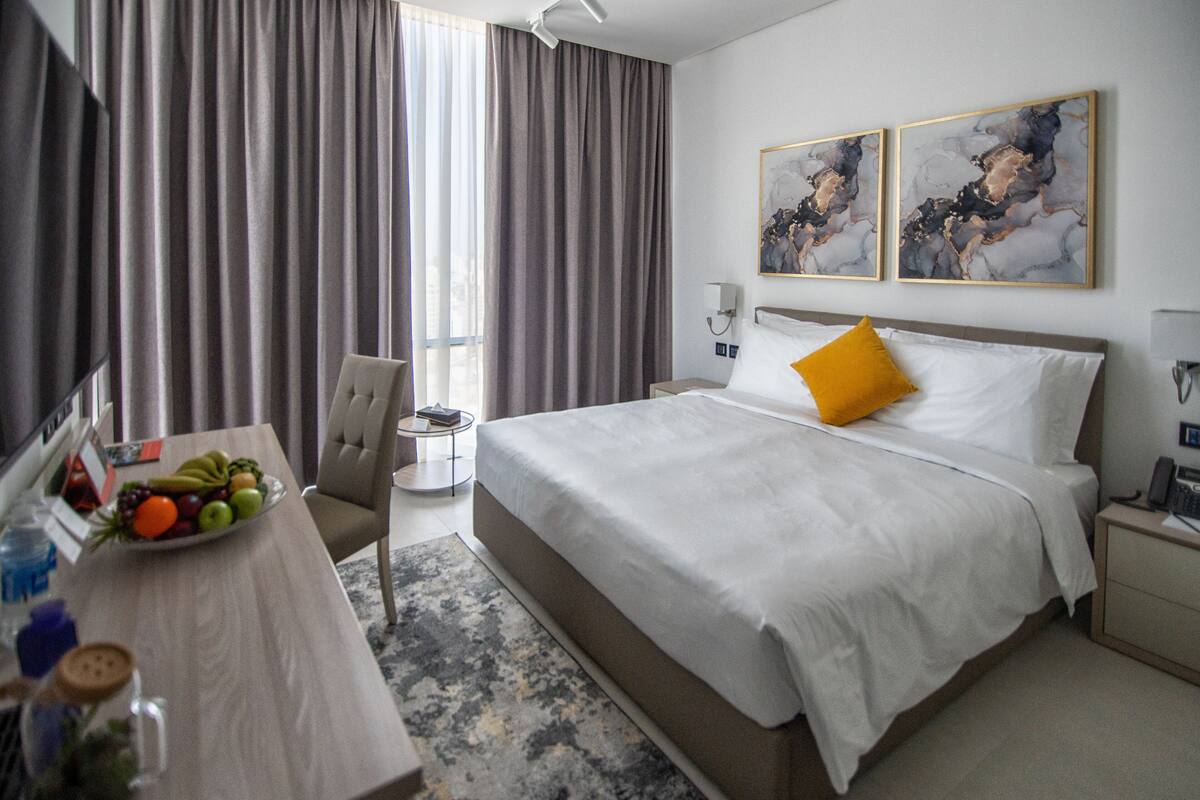 A hotel room with a bed, a desk with a fruit bowl, a chair, a nightstand, and two abstract paintings on the wall.