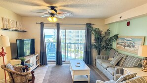 A living room with a TV, a ceiling fan, a sofa, a chair, and a large window with curtains. - Beautiful Ocean Views 2/2 Condo  (Orange Beach)