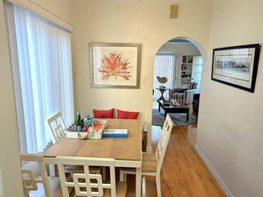 Ein Essbereich mit Holztisch und weißen Stühlen, ein gerahmtes Kunstwerk darüber und Blick in ein Wohnzimmer durch einen Bogen.