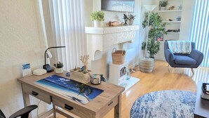 A modern living room with a wooden table, a blue armchair, and a round blue rug. There are shelves with plants and decorative items, and a window with blinds.