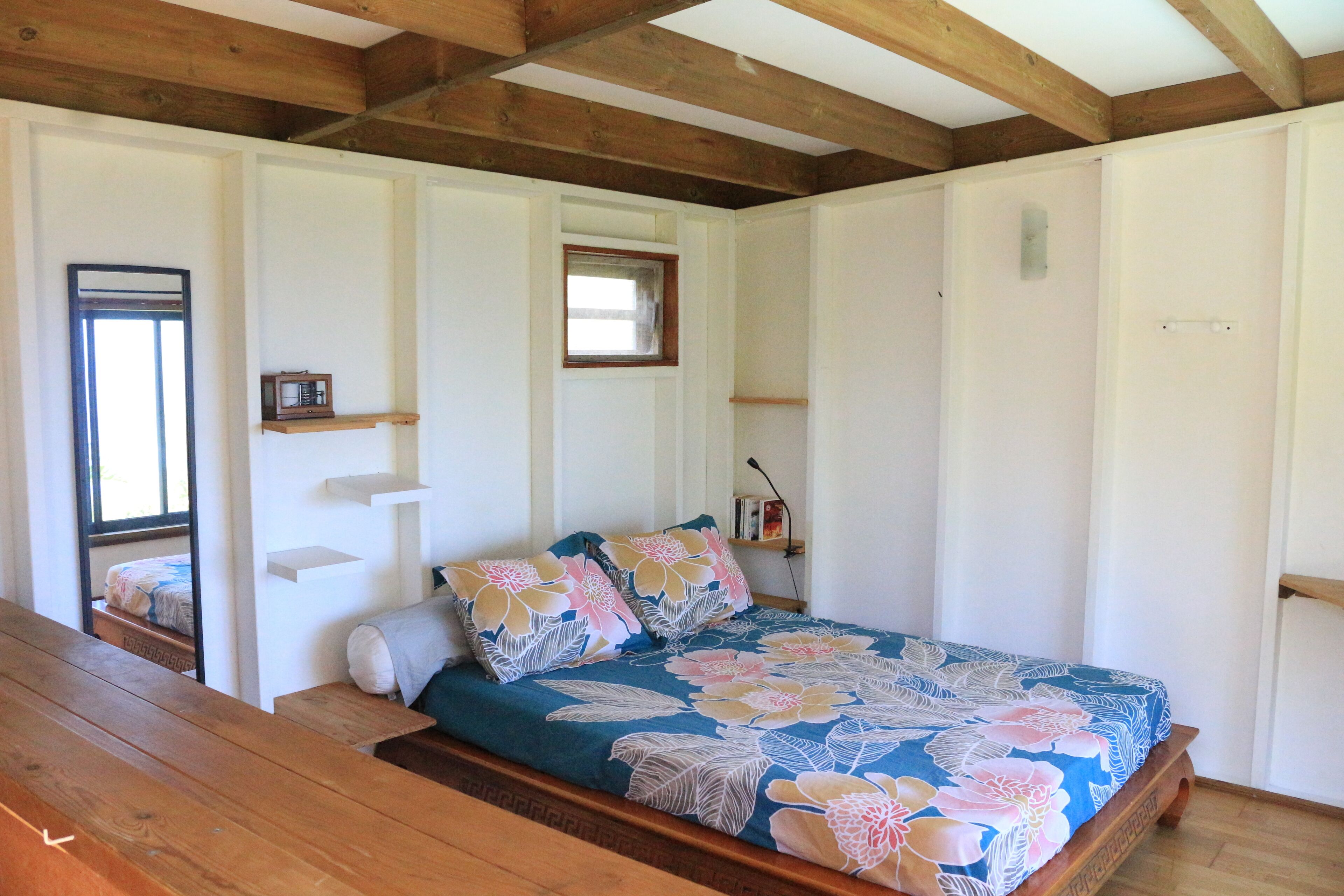 A bedroom with a wooden bed, floral bedding, a small window, and a mirror on the wall.