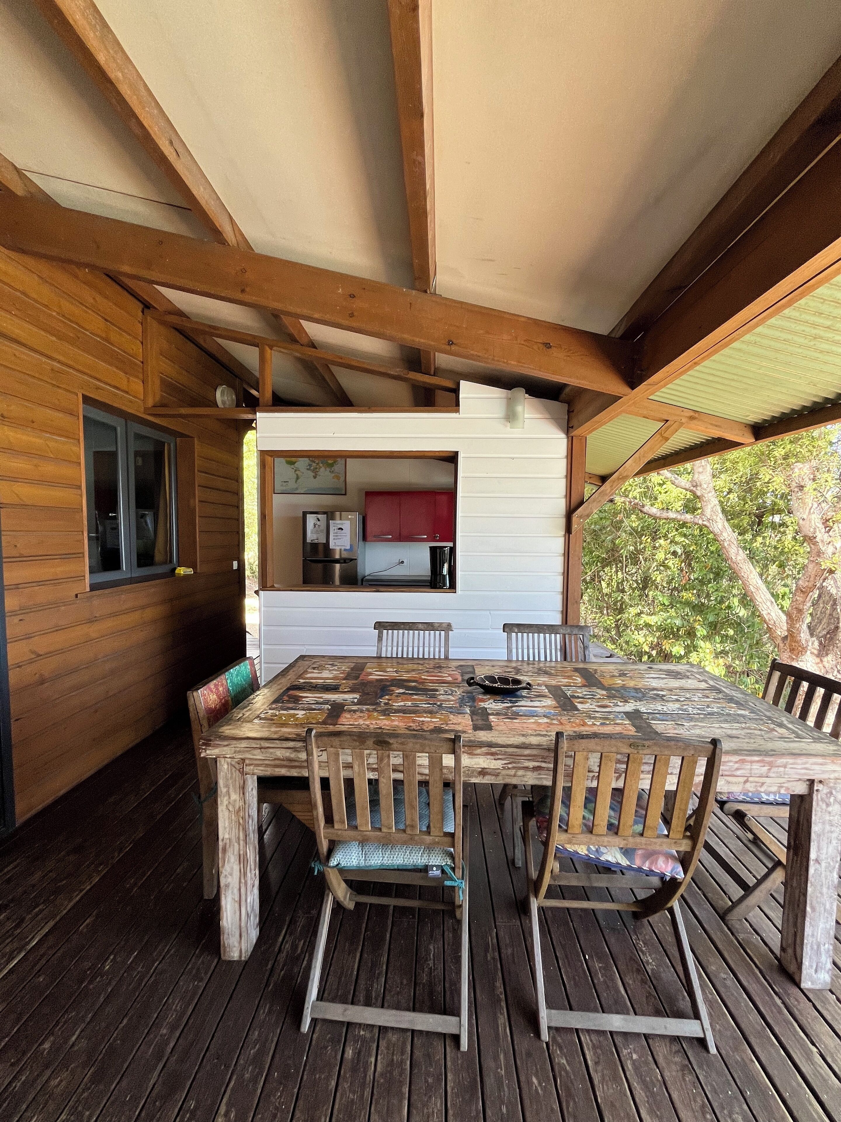 Une terrasse en bois avec une table et des chaises rustiques, un coin cuisine avec des meubles rouges et une vue sur les arbres.