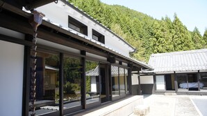 Een traditioneel Japans gebouw met grote schuiframen, een houten terras en uitzicht op een bebost heuvellandschap.