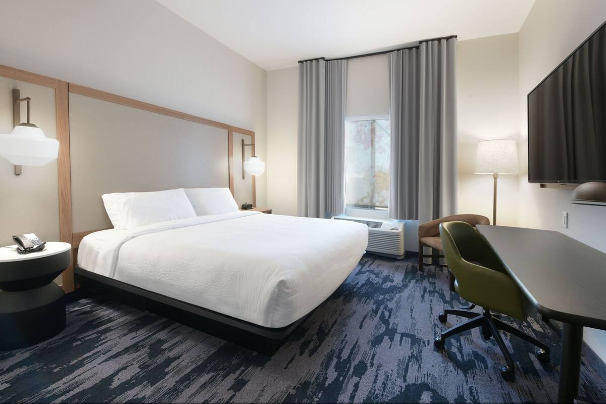 Een hotelkamer met een groot bed, een bureau, een stoel, een televisie, een lamp en een raam met gordijnen.