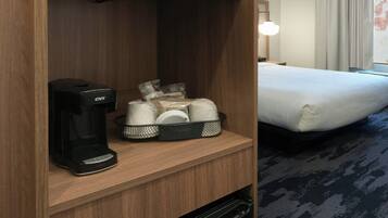 Habitación de hotel con un mueble de madera que contiene un microondas, una cafetera y un refrigerador pequeño. Hay una cama con colchón blanco y una ventana con cortinas.