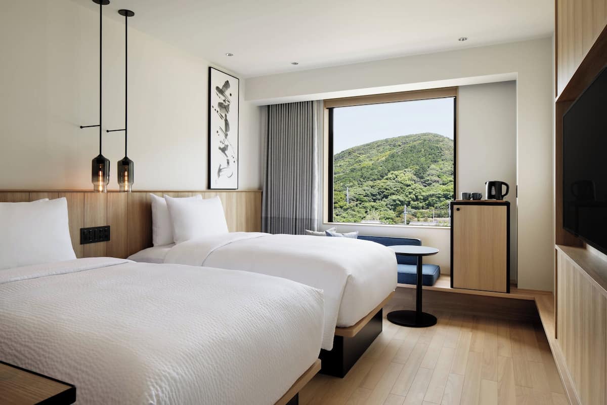 Een hotelkamer met twee bedden, een groot raam met uitzicht op een groene heuvel, een kleine tafel en een televisie.