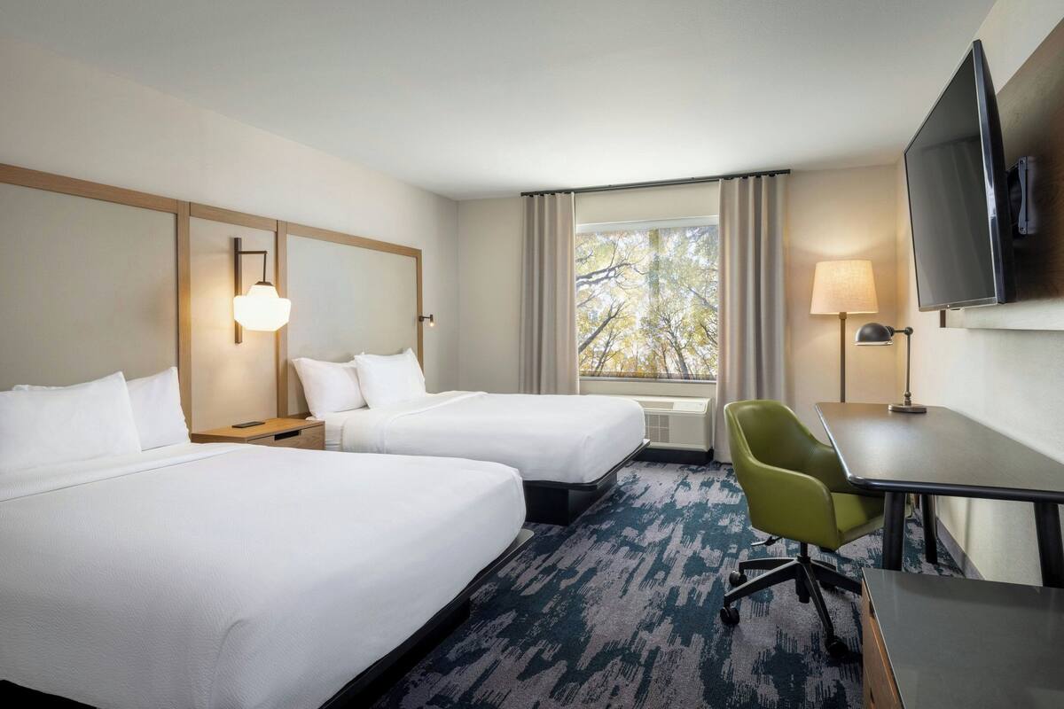A hotel room with two beds, a desk, a TV, and a window with curtains.