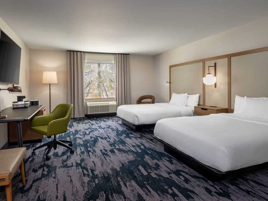 A hotel room with two beds, a desk with a chair, a television, a window with curtains, and a lamp.