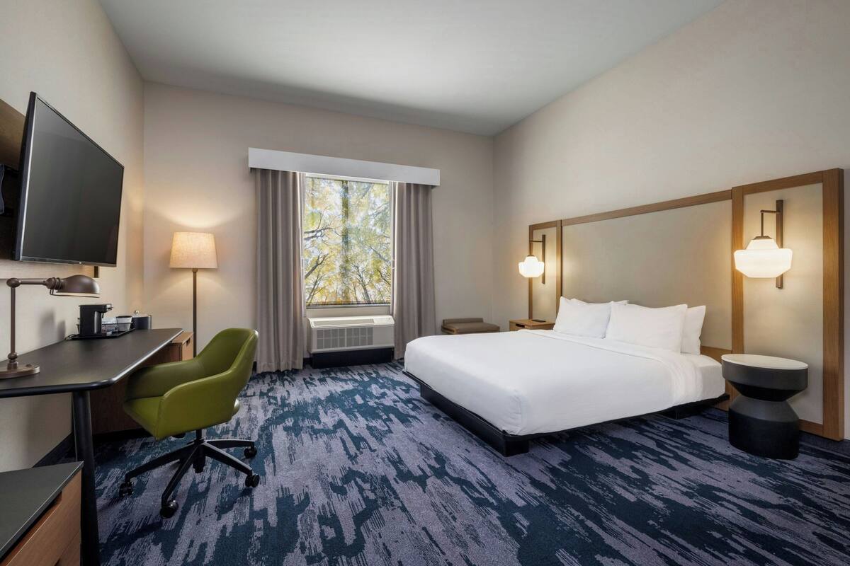 A hotel room with a bed, desk, chair, TV, lamp, and window with curtains.