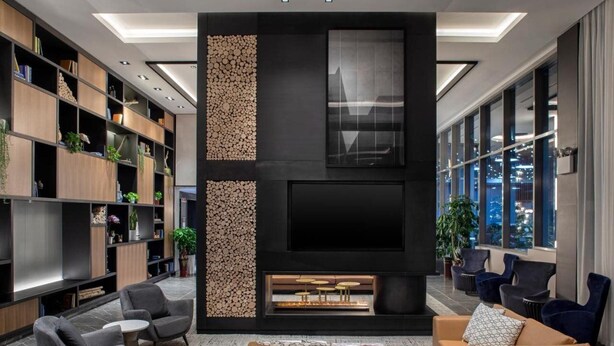 A modern living room with a fireplace, comfortable seating, and a large window offering a city view. - Four Points by Sheraton Flushing (Flushing)