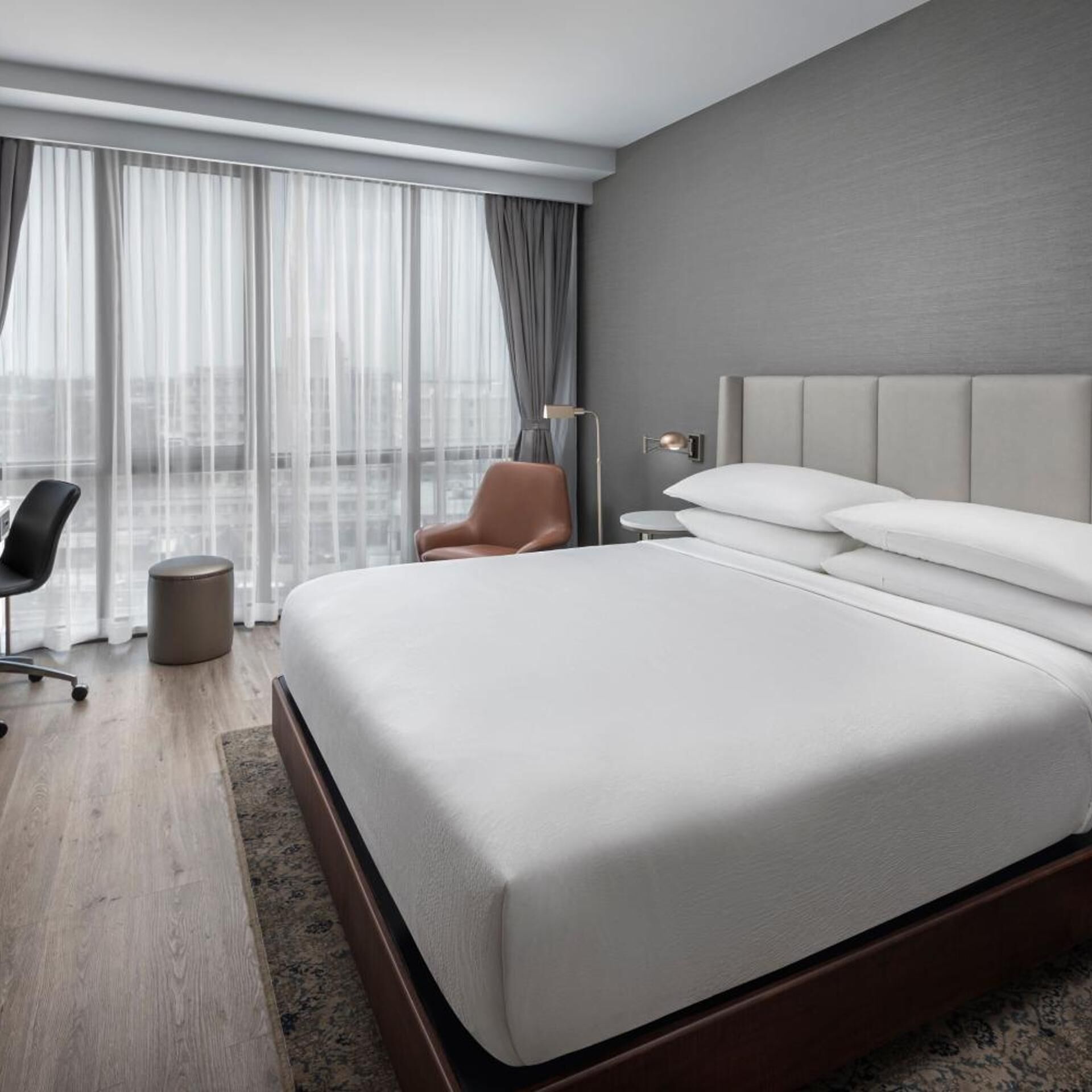 A modern hotel room with a large bed, a chair, a bedside table, and a view of the city through sheer curtains.