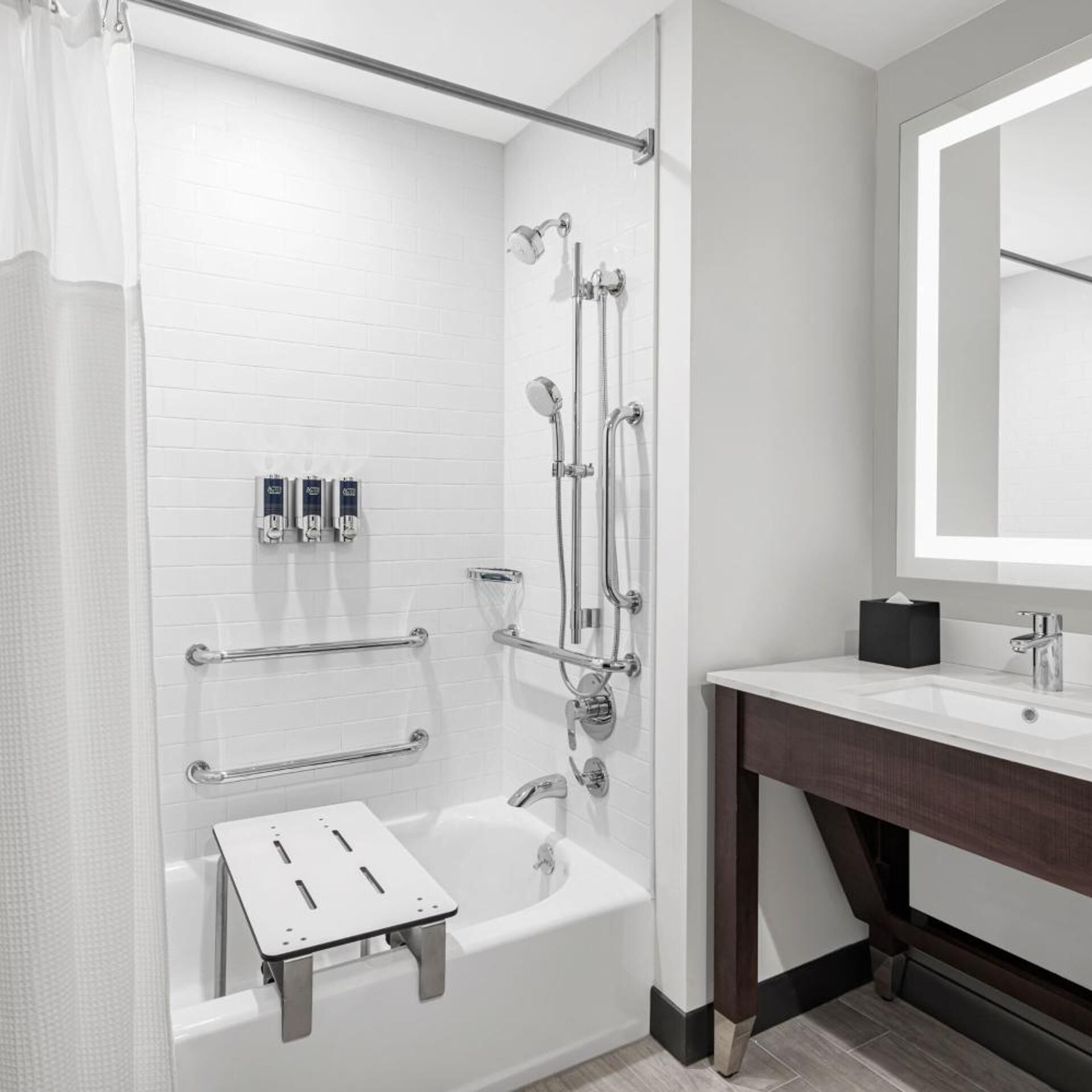 A modern bathroom with a white bathtub, a shower with multiple heads, a handrail, and a vanity with a mirror.