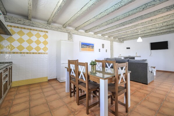 A kitchen with a dining table and chairs, a refrigerator, and a TV mounted on the wall.