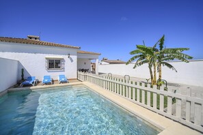 A swimming pool with blue lounge chairs, a white fence, and a palm tree.