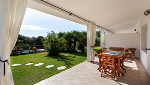 Un porche cubierto con una mesa y sillas, vista a un jardín y árboles, y una piscina al fondo.