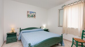 Un dormitorio con una cama, dos mesitas de noche con lámparas, una silla, una ventana con cortina y un cuadro colgado en la pared.