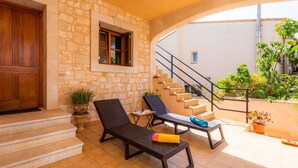 A stone-walled room with a wooden door, a window with shutters, a staircase, and two sun loungers with towels.