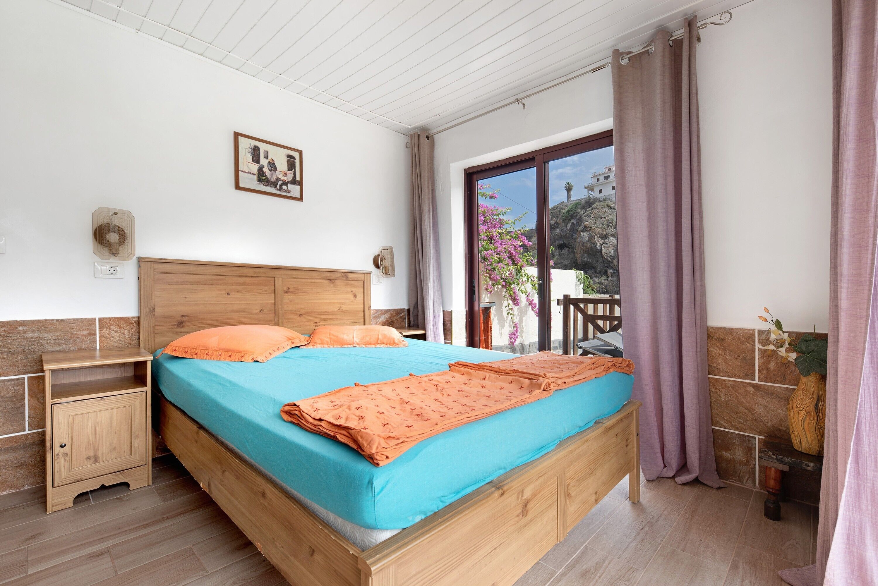 Une chambre à coucher avec un lit en bois, une table de chevet, une fenêtre avec des rideaux, une lampe fixée au mur et une petite table sur laquelle est posé un vase.