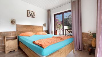 Une chambre à coucher avec un lit en bois, une table de chevet, une fenêtre avec des rideaux, une lampe fixée au mur et une petite table sur laquelle est posé un vase.