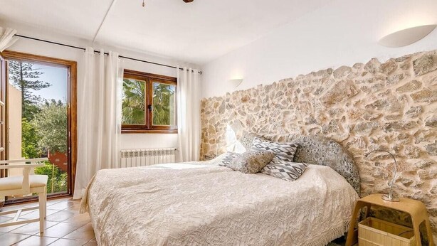 Une chambre avec un mur en pierre, un lit avec une literie à motifs, une table de chevet en bois et une vue à travers une porte vitrée.