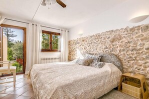 Une chambre avec un mur en pierre, un lit avec une literie à motifs, une table de chevet en bois et une vue à travers une porte vitrée.