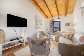 Een woonkamer met een flatscreen televisie, een houten plafond, een witte bank, een ronde glazen tafel en een witte stoel met een gebreide rugleuning.