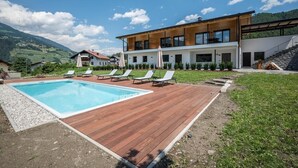 Un edificio moderno con piscina, sillas de descanso y montañas al fondo.