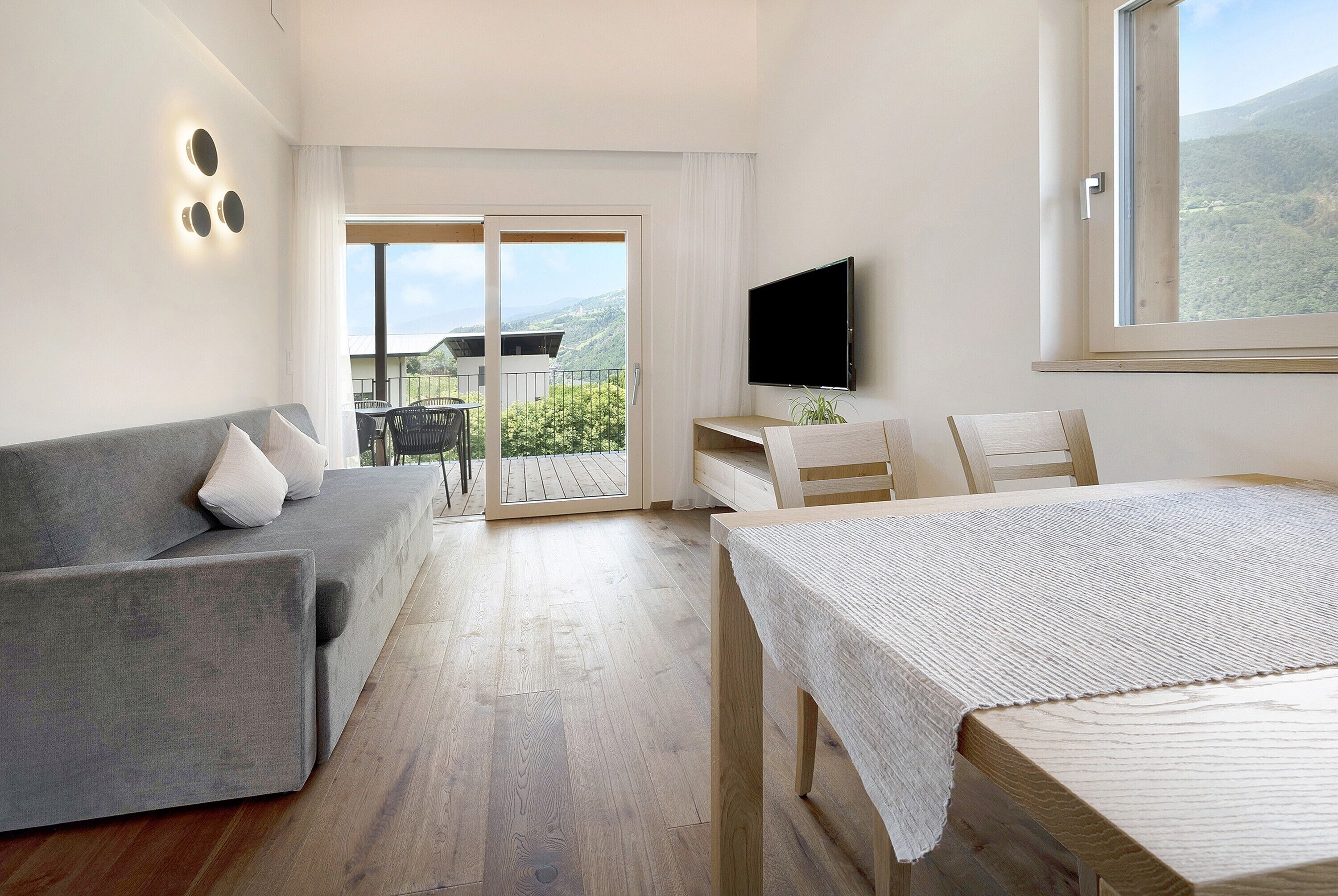 Una camera d'hotel moderna con un divano grigio, un tavolo in legno, una TV e un balcone con vista sulle montagne.