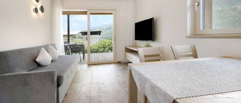 Una camera d'hotel moderna con un divano grigio, un tavolo in legno, una TV e un balcone con vista sulle montagne.
