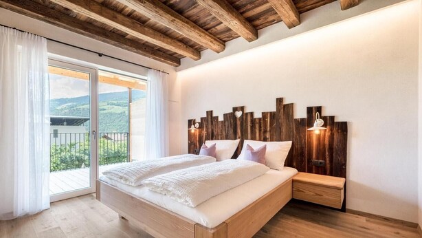A bedroom with a wooden headboard, a large bed, a nightstand, and a sliding glass door to a balcony. - Apartment 'Sommersbergerhof' with Mountain View, Shared Pool and Wi-Fi (Brixen)