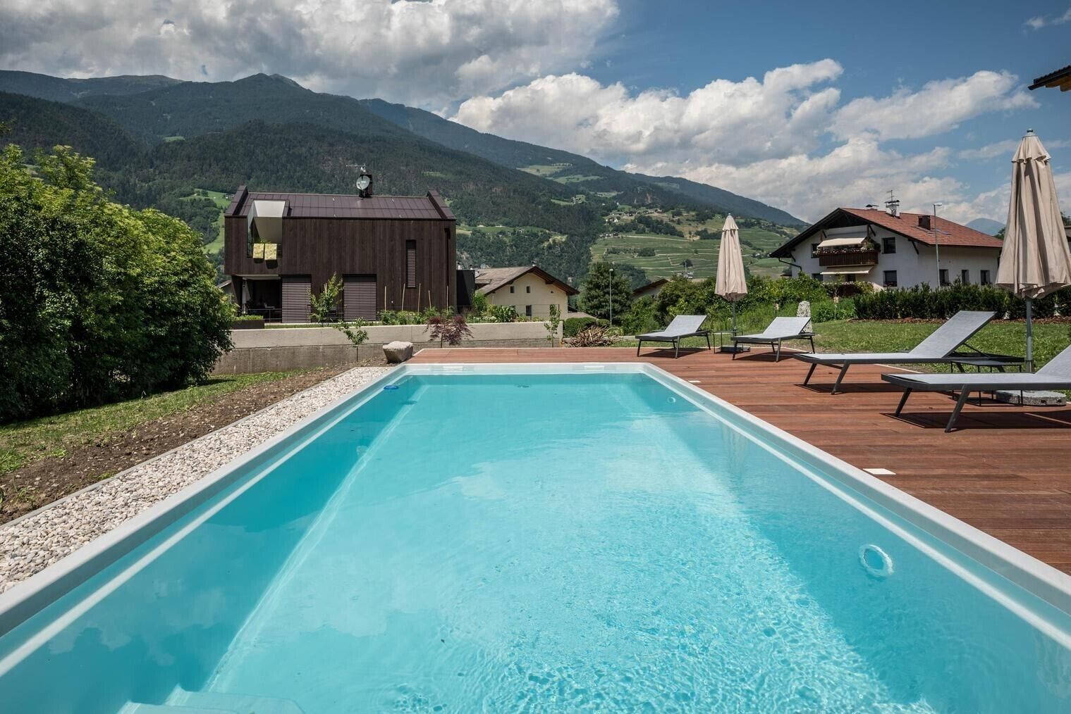 Una piscina con sillas de descanso y sombrillas, rodeada de casas y montañas.