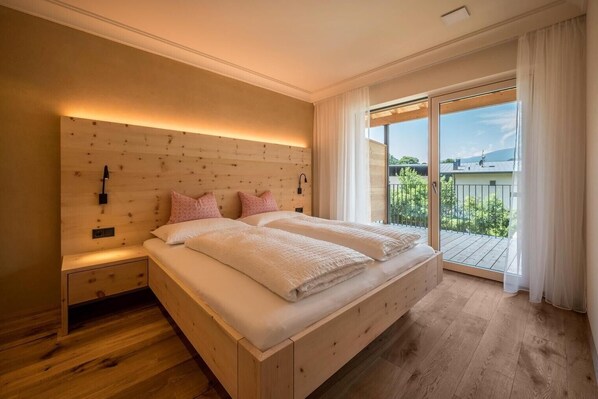 Une chambre à coucher avec une tête de lit en bois, un grand lit avec une literie blanche, une table de chevet et une porte-fenêtre coulissante donnant sur un balcon.