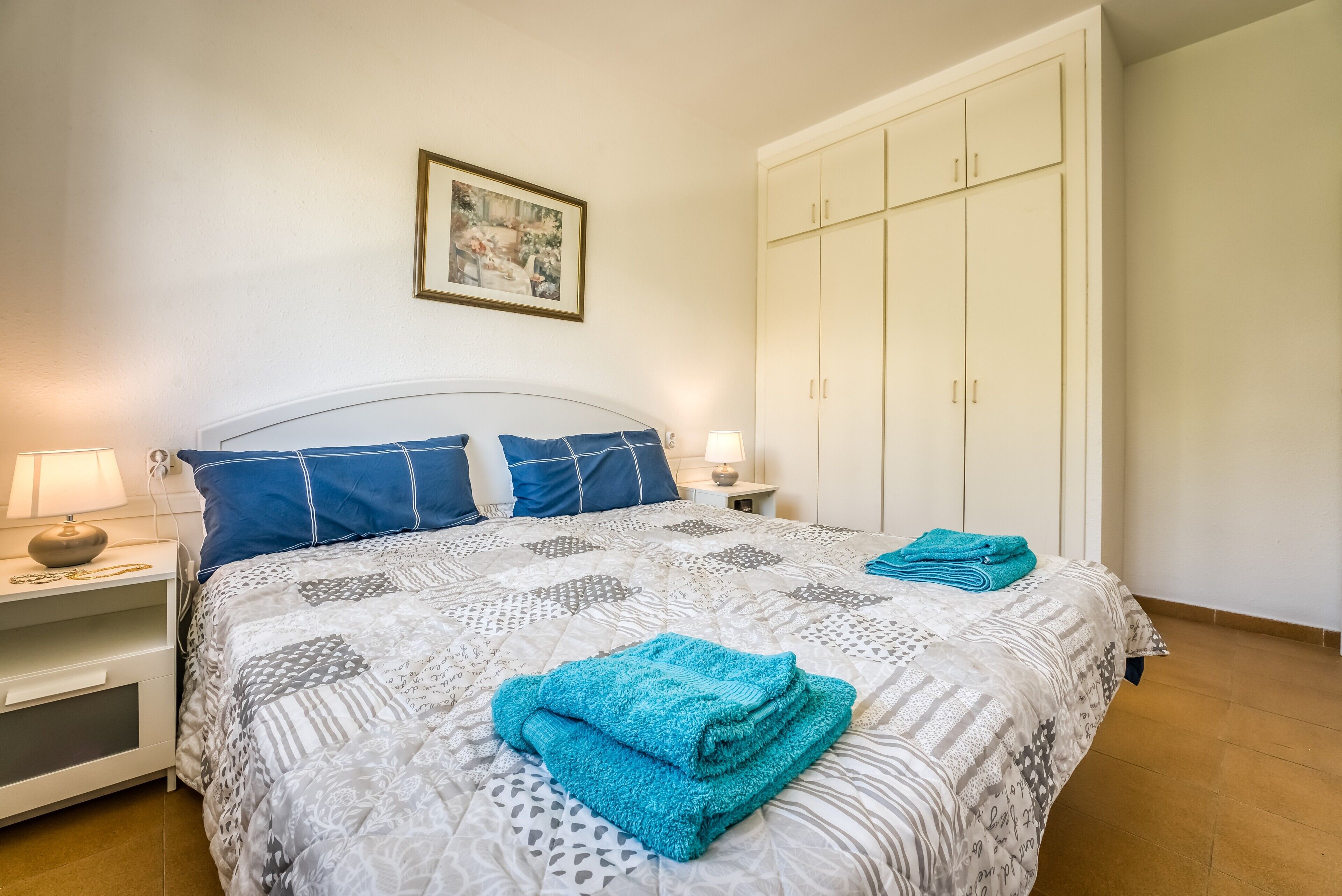 Una cama bien hecha con ropa de cama estampada, una lámpara de noche y una toalla doblada sobre la cama.