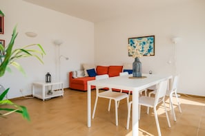 Un salon moderne avec une table à manger blanche, des chaises blanches, un canapé rouge agrémenté de coussins bleus, un lampadaire blanc et un tableau accroché au mur.