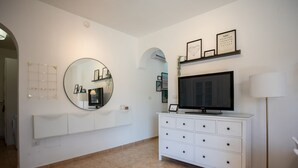 Ein modernes Wohnzimmer mit einem großen Flachbildfernseher auf einem weißen Schrank, einem runden Spiegel an der Wand und einem kleinen Regal mit gerahmten Bildern.