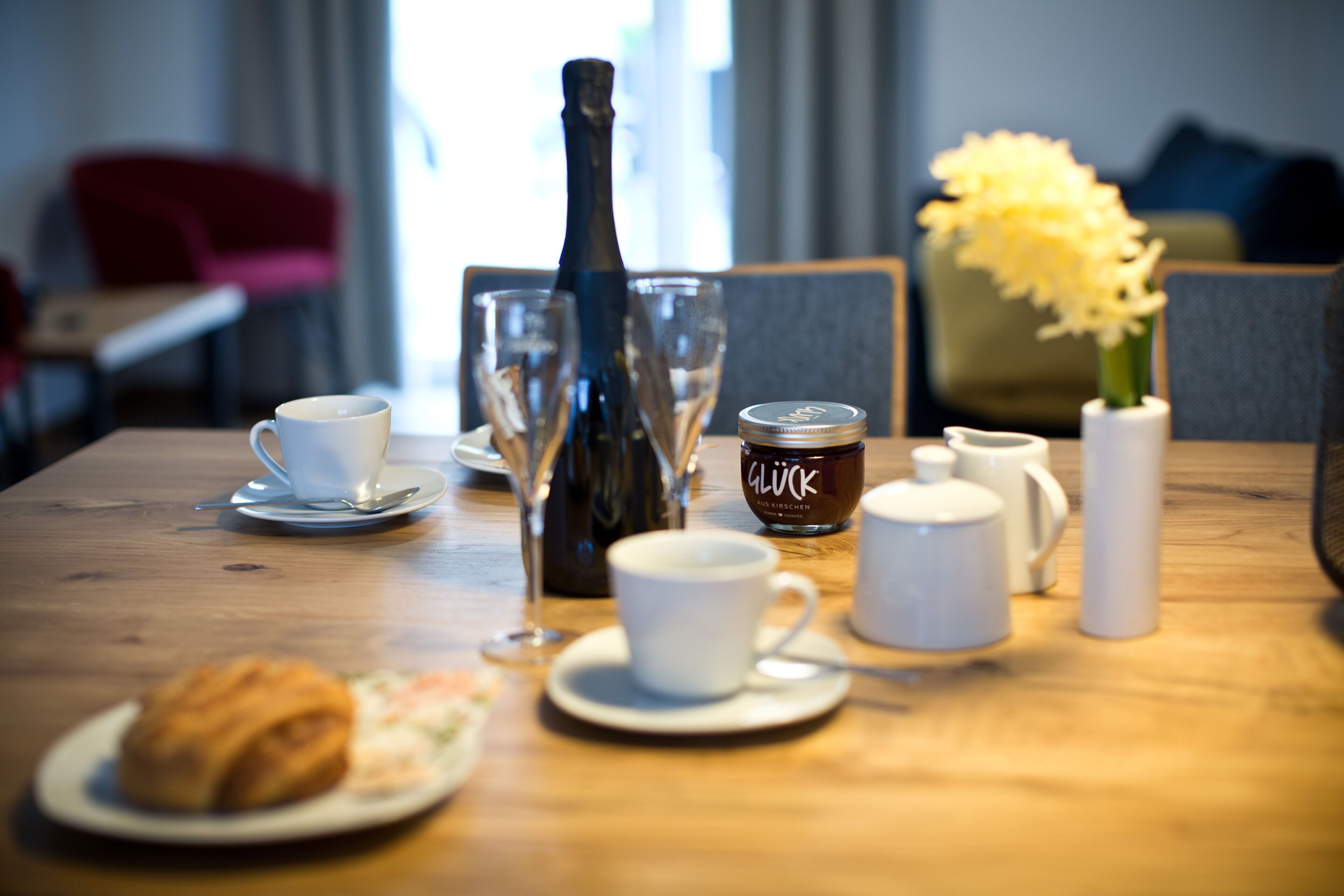 Une table à manger avec une bouteille, des verres, un bocal, un vase avec des fleurs, une assiette avec une pâtisserie et une tasse.