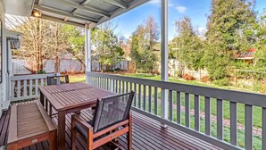 A covered outdoor patio with a wooden table and chairs, a white railing, and a view of a backyard with trees and a fence.