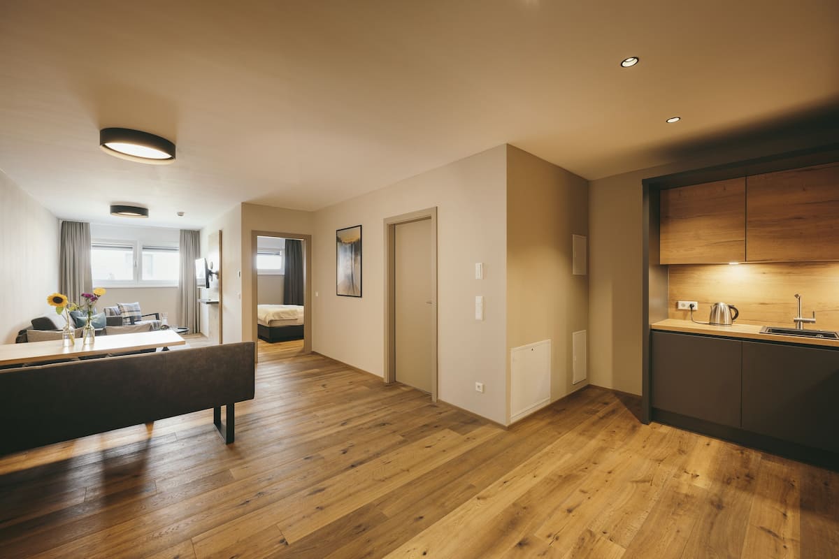 A modern apartment with a wooden floor, a kitchen with a sink and kettle, a dining area with a bench, and a living room with a sofa and curtains.