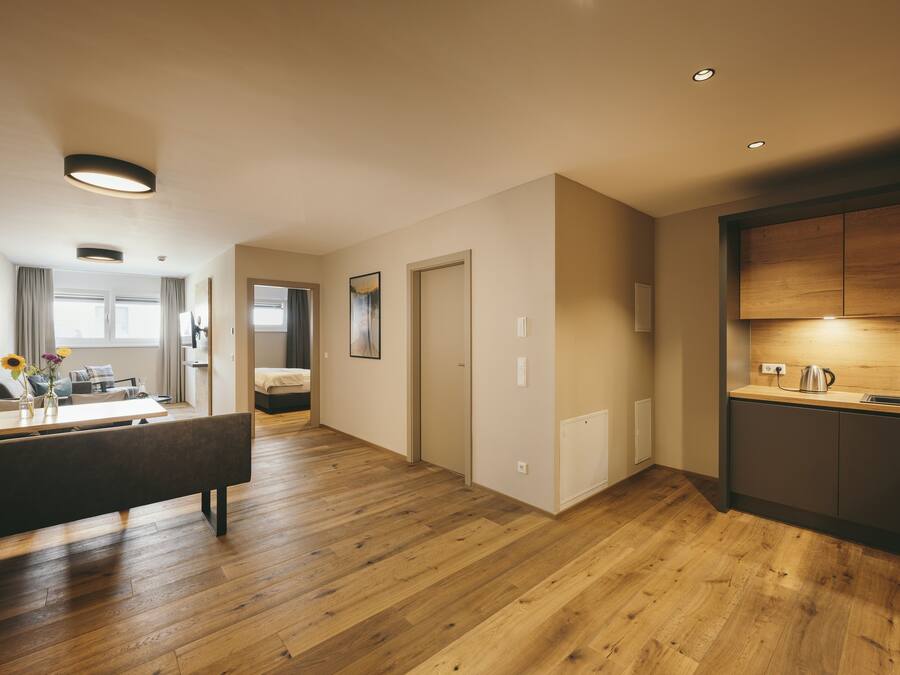 Un apartamento moderno con suelo de madera, una cocina con lavaplatos y hervidor, una zona de comedor con banco, y una sala de estar con sofá y cortinas.