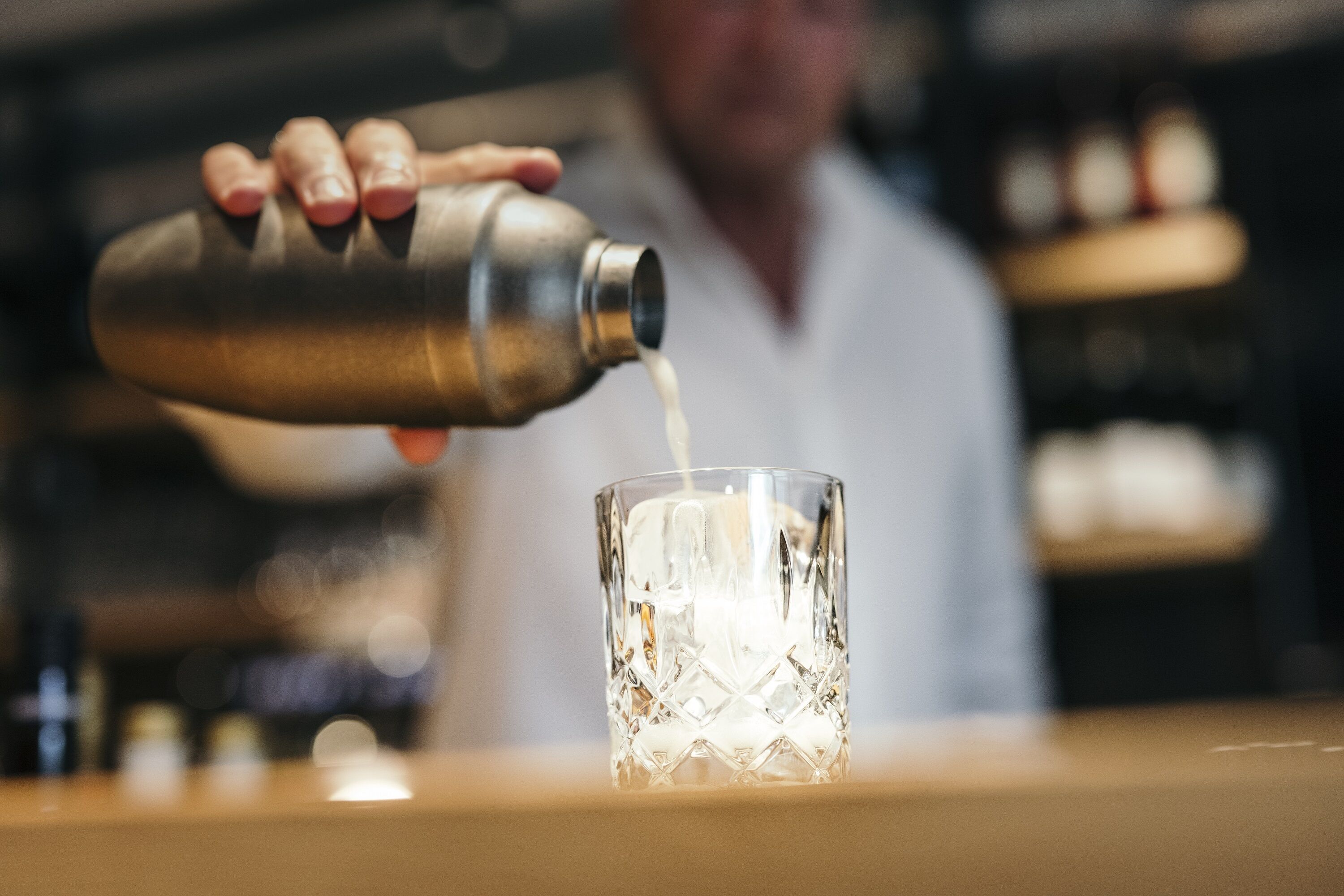A person pouring a liquid from a metal shaker into a clear glass.