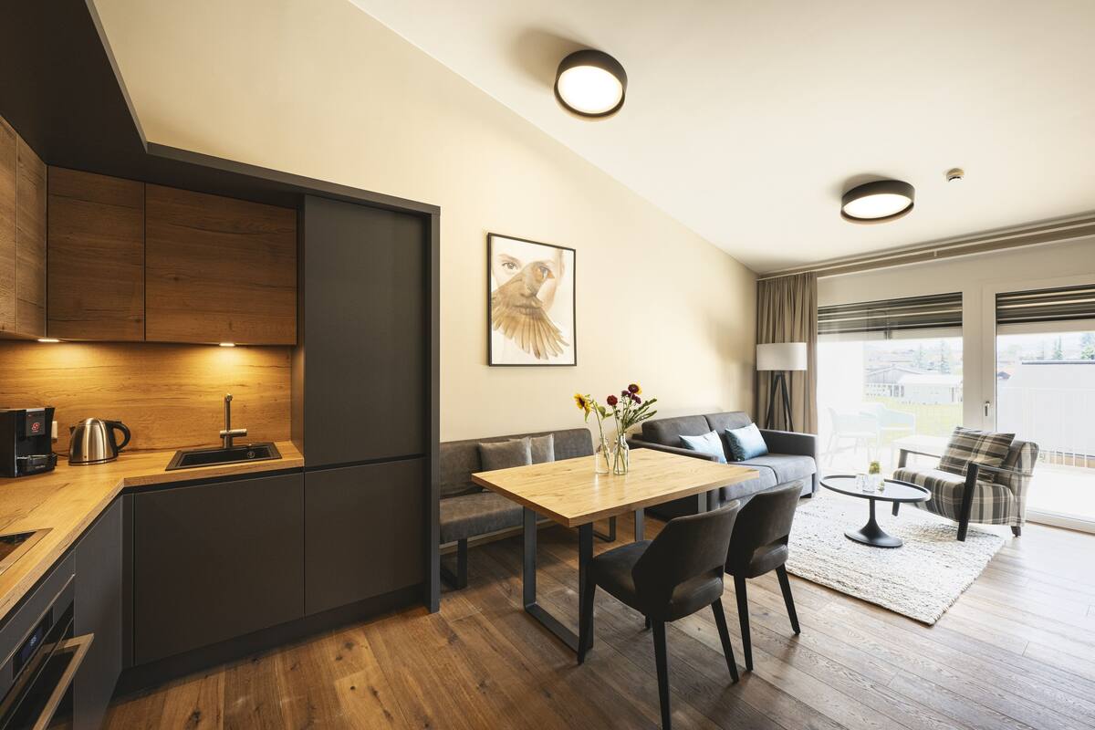A modern kitchen with a built-in oven, a dining area with a wooden table and chairs, a sofa, and a large window offering a view of the outdoors.