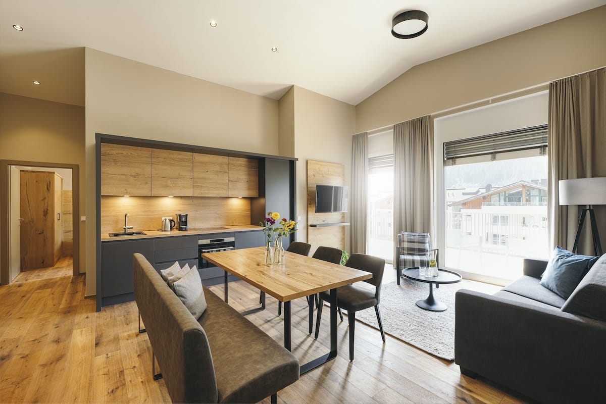A modern living room with a wooden dining table, grey sofa, and a large window with curtains.