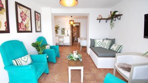 A living room with turquoise sofas, a white coffee table, and a grey sofa with patterned cushions. There are framed floral artworks on the wall, a hanging light fixture, and a bookshelf with books and plants.