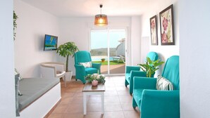 A living room with teal armchairs, a white coffee table, and a TV on the wall. There is a plant and a view of the outdoors through a glass door.