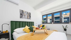 A modern hotel room with a green upholstered headboard, a bed with white and yellow bedding, a bedside table with a lamp, a small plant, and a window with a view of buildings. - Apartment Skyline (Konavle)