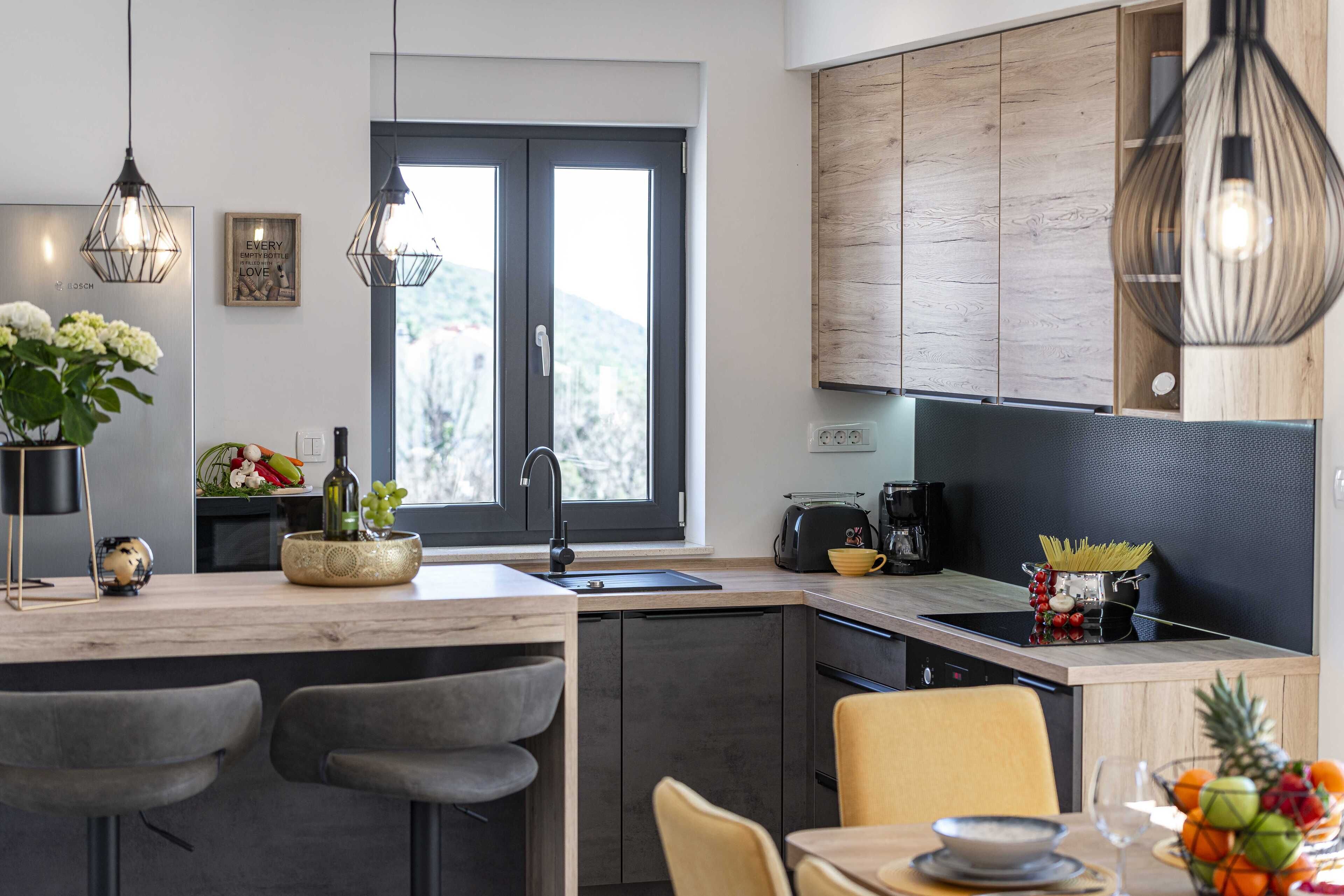 A modern kitchen with a central island, bar stools, a dining table, and a view of the outdoors through a large window.