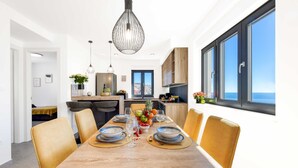 A modern dining area with a wooden table set for four, surrounded by yellow chairs, a fruit bowl, and a view of the sea through large windows. - Apartment Skyline (Konavle)