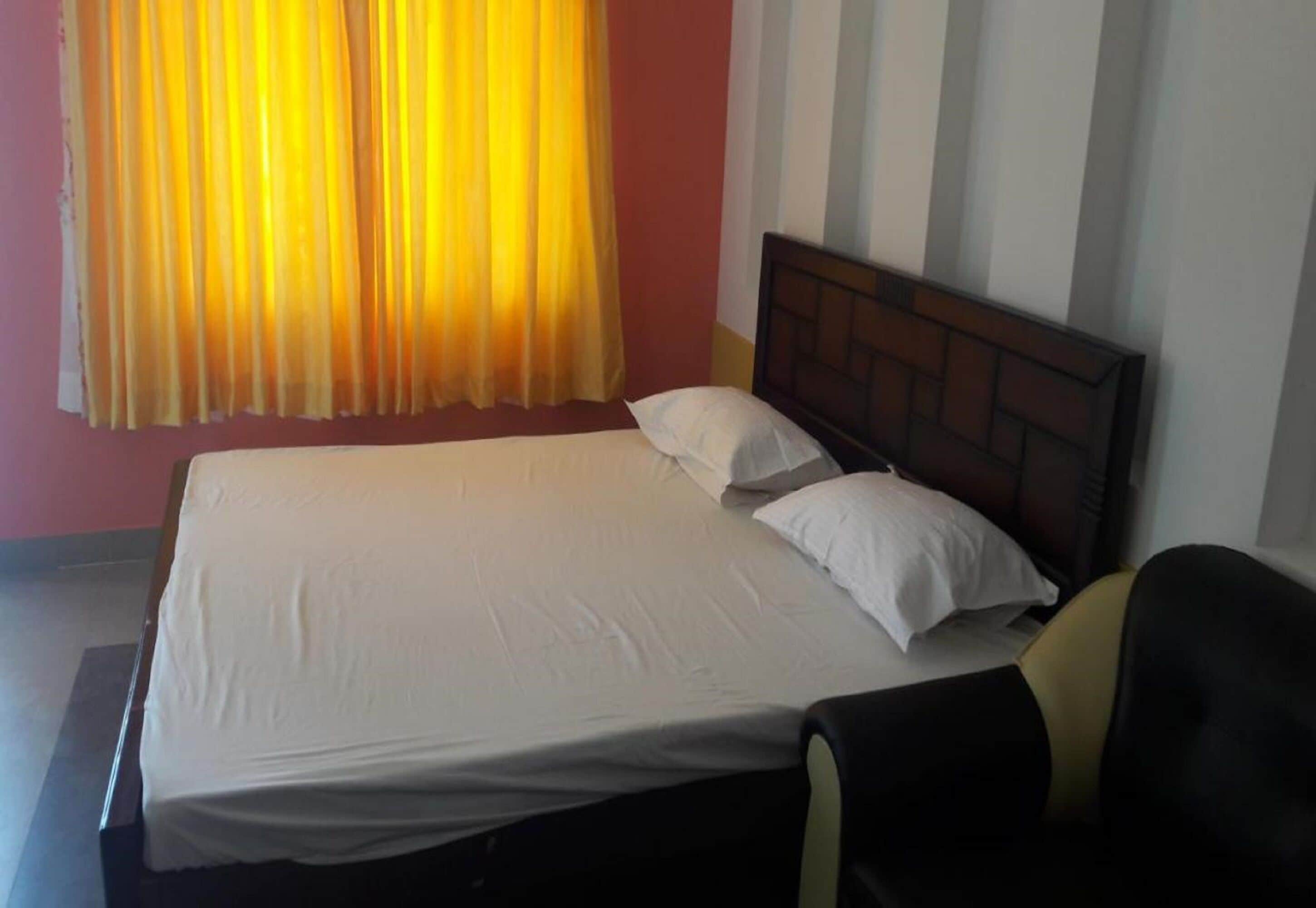 A single bed with white linens and a wooden headboard in a room with a plain wall and a window with yellow curtains.